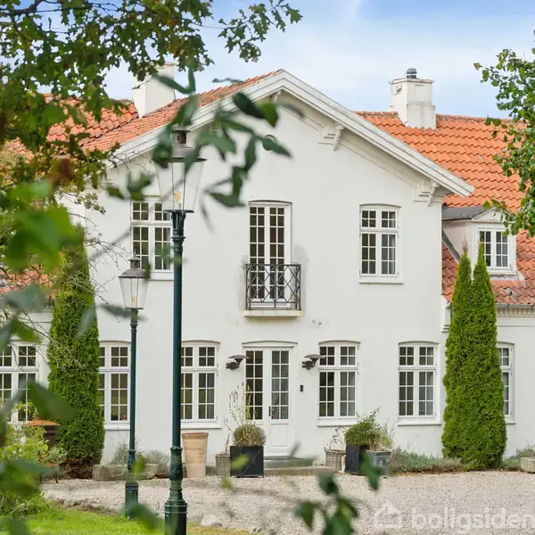 Hvid bygning med rødt tegltag står omgivet af høje træer og en grusbelagt gårdsplads. Gadelamper og planter dekorerer området, mens klare blå himmel ses i baggrunden.