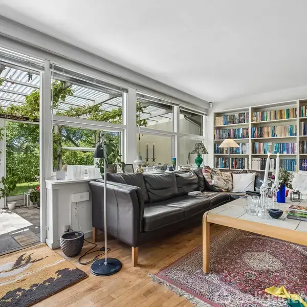 En sort sofa står foran en stor reol fyldt med bøger, mens to lamper lyser rummets hjørner. Store vinduer og en glasdør afslører en grøn, frodig have udenfor.