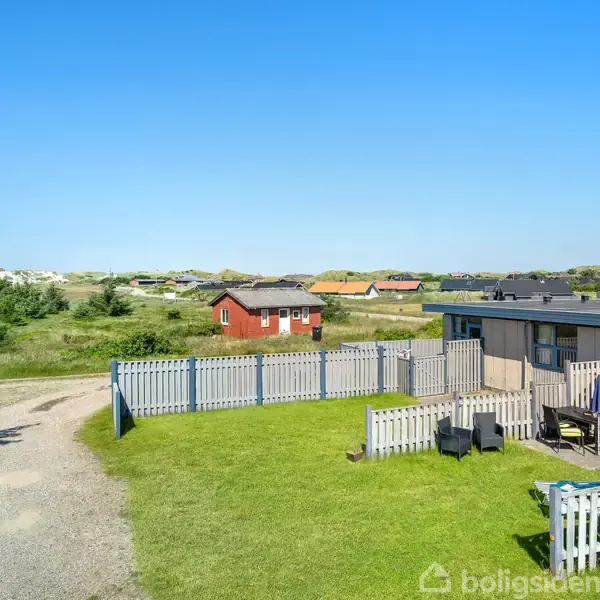 Et sommerhus med terrasse, omgivet af et hvidt stakit. I baggrunden ses en rød hytte og åbne grønne marker med spredte træer under en klar blå himmel.