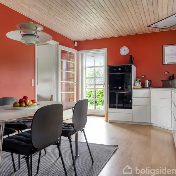 Et køkken med orange vægge. Et bord med sorte stole og en frugtskål. Hvide skabe langs væggen. En glasdør fører ud til en grøn have. En loftslampe hænger over bordet.
