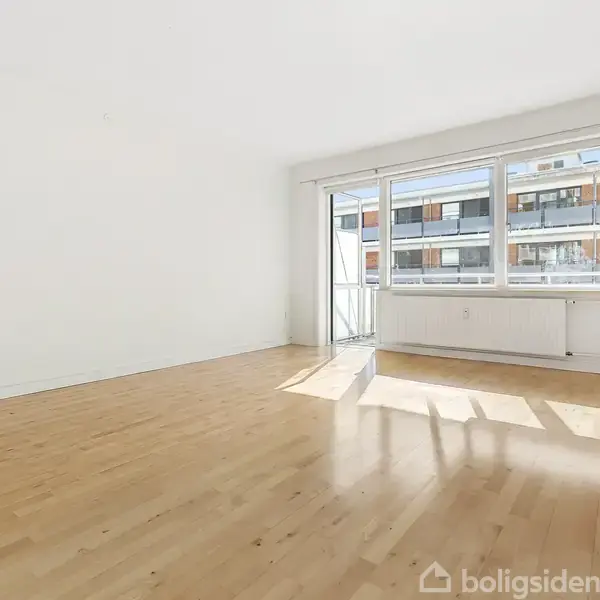 Et tomt rum med trægulv, store vinduer og en dør til en balkon, der slipper lys ind. Udsigt til en bygning udenfor gennem vinduerne.