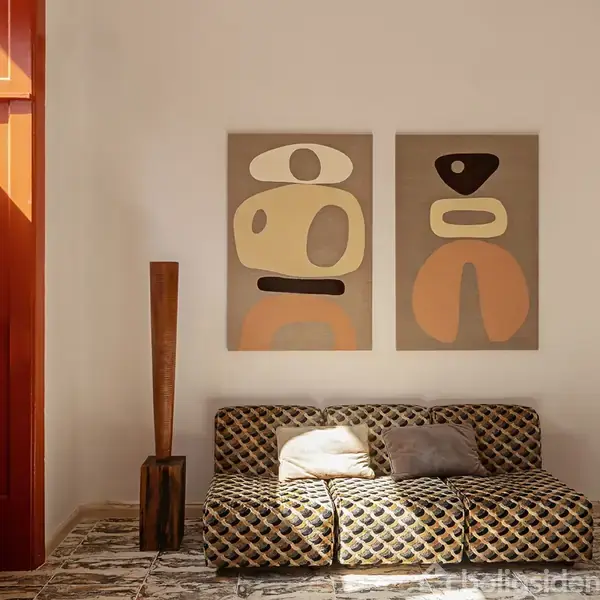 Sofa med geometrisk mønster står mod lys væg med to abstrakte malerier. Til venstre står en vase med solsikker, mens en høj, skulpturel lampe står til højre. Sollys strømmer ind.