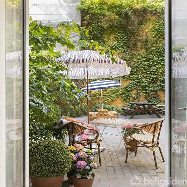 En terrasse med to flettede stole og et lille, rundt bord under en stribet parasol. Baggrunden har en gul mur dækket af grønne planter og en træbænk.