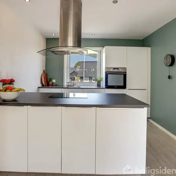 En hvid køkkenø med sort bordplade står centralt. Et moderne emfang hænger over den. I baggrunden er der et vindue, ovn og skabe. Væggen er malet grøn.