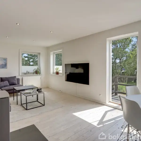 Stue med sofa og sofabord; vægmonteret TV; lyst, enkelt design; store vinduer med udsigt til grønt område; spisebord til højre; solrig atmosfære.