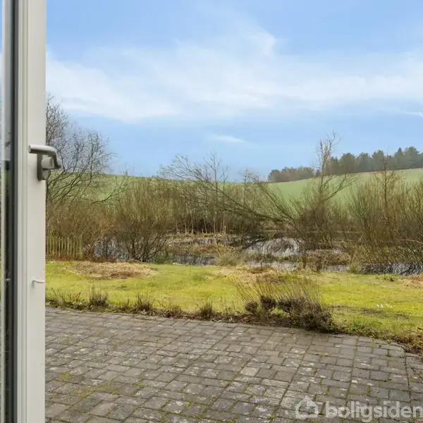 En glasdør fører ud til en flisebelagt terrasse med udsigt over en græsplæne og en lille sø, omgivet af træer og et bakket landskab i baggrunden under en blå himmel.