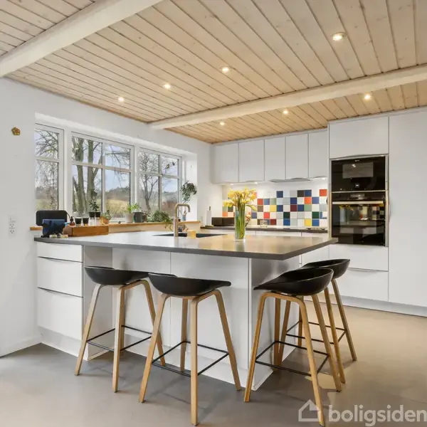 Et moderne køkken med en central ø omgivet af tre sorte barstole. Klinkegulv og hvidt loft med spots. Store vinduer giver udsigt til en have med grønne planter. Farverigt flisebagsæt ved komfuret.