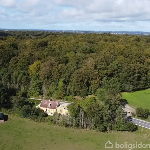 Et hus står stille omgivet af grønne marker og tæt skov, mens en vej løber forbi. Himlen er delvist overskyet.