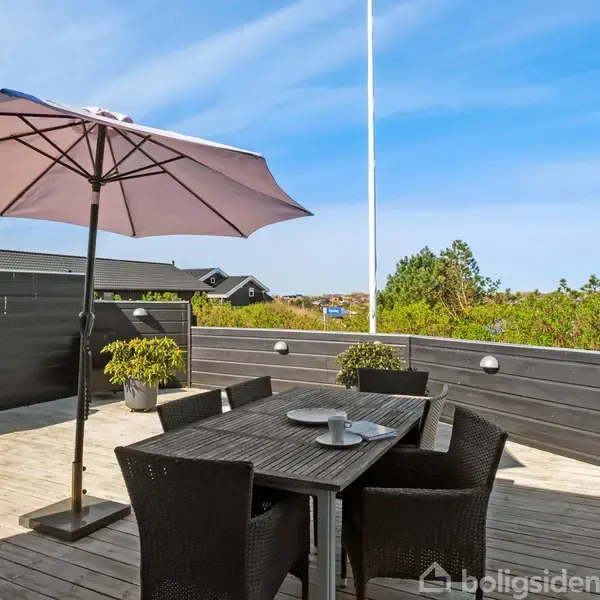 En stor parasol skygger over et udendørs spisebord med fire stole. Bordet står på en terrasse med træbelægning, omgivet af hæk og grill i baggrunden.