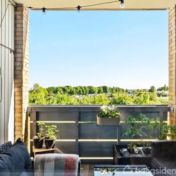 Altan med sofagruppe og planter står stille foran en udsigt over grønne træer og blå himmel.