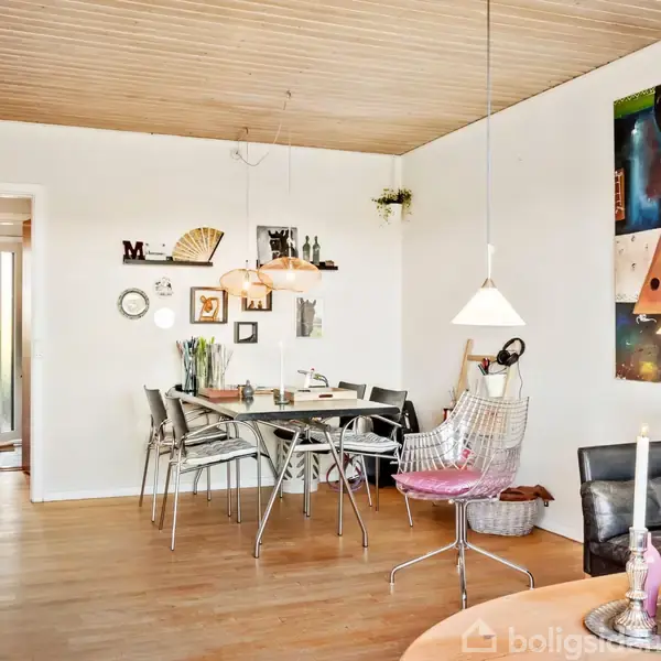 Spisestue med et bord omgivet af stole, udsmykket med moderne kunst på væggen. Lyse lamper hænger fra træloftet, og rummet har trægulv. En åbning fører til et andet område.