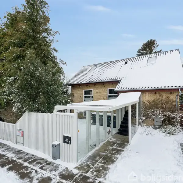 Et hus dækket af et lag sne står i en snedækket have. Huset har et skråt tag og en tilstødende opbevaringsstruktur. Sne beklæder også den omkringliggende indkørsel.