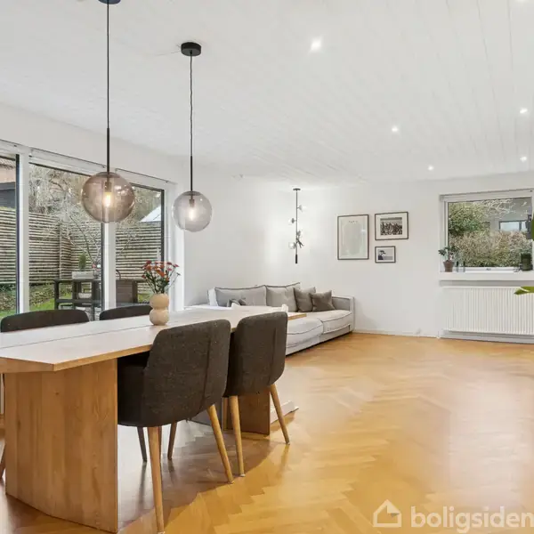 Spisebord omgivet af fire stole under to hængende lamper, i en stue med parketgulv og en grå sofa. Store glasdøre åbner ud til en have, mens hylder og vægkunst dekorerer rummet.