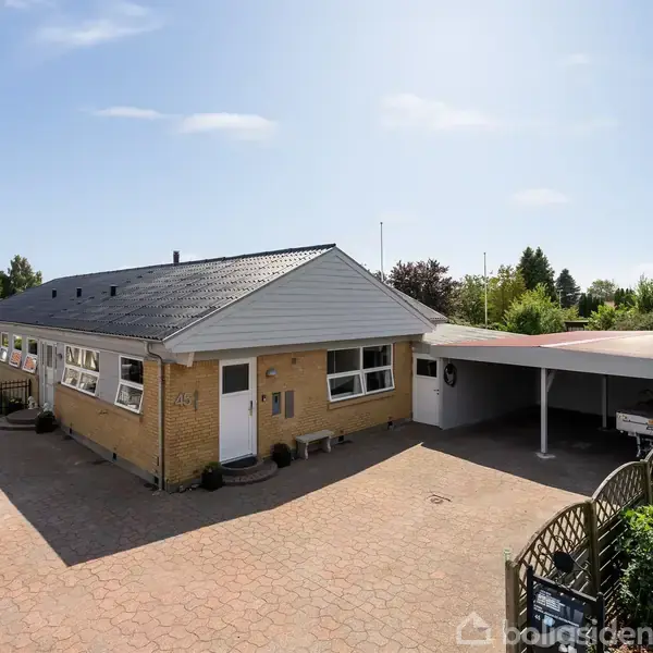 Et enfamiliehus af mursten står stille ved siden af en stor, flisebelagt indkørsel. Huset har en carport og er omgivet af en have med hække og buske under en blå himmel.