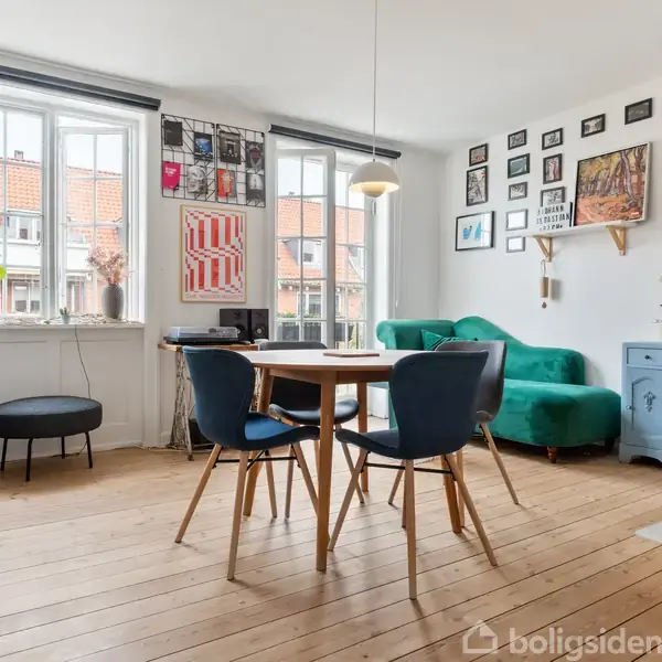 Stue med rundt spisebord og blå stole står på trægulv. Grøn sofa ved væggen med mange kunstværker. Store vinduer lader lys strømme ind, med planter på vindueskarmen.