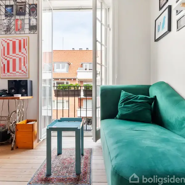 Grøn sofa står i lys stue ved fransk altan. Bord ved siden og plakat med tekst "CARL NIELSEN MUSEET". Bygninger kan ses udenfor, giver en bymæssig atmosfære.