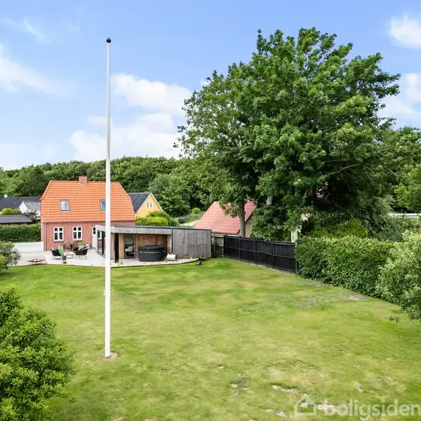 Et rødt murstenshus med oranger tag står i en grøn have med græsplæne. Flagstang rejser sig tæt på huset, og omgivelserne inkluderer træer og buske under en blå himmel.
