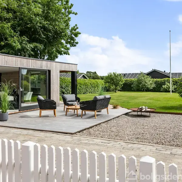 Rødt murstenshus med orange tegltag står på flisebelagt gårdsplads. Havemøbler står foran huset. I baggrunden er der grønne hække og træer under en blå himmel med skyer.