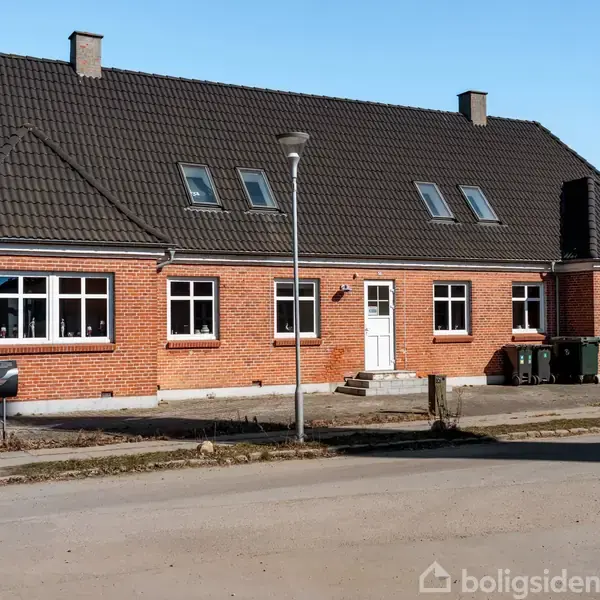 Et rødt murstenshus med mørkt tag har flere vinduer og en hvid dør. Det står ved en vej, omgivet af en lygtepæl og en skraldespand.