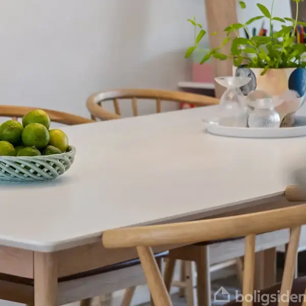 En hvid spisebordsoverflade har en skål med grønne limes. På bordet står også en potteplante ved siden af en bakke med glas. Træstole omgiver bordet.