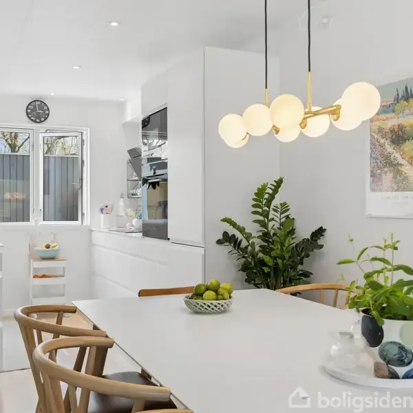 Et lyst køkken/alrum med et hvidt bord i forgrunden dekoreret med planter og frugter. Bagved ses lyse køkkenelementer, ovn og en vask. To billeder hænger på væggen.