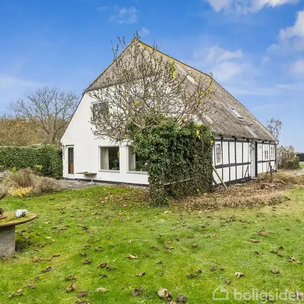 Hus med hvid facade og stråtag står i en frodig have med græsplæne og træer. Et lille rundt bord med en dekoration står til venstre. himlen er blå med skyer.