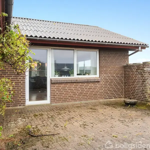 Røde murstensvægge med store vinduer åbner ud til en flisebelagt terrasse. Ranken til venstre vokser op ad væggen. Murstensvæggen omkranser en lille gårdhave.