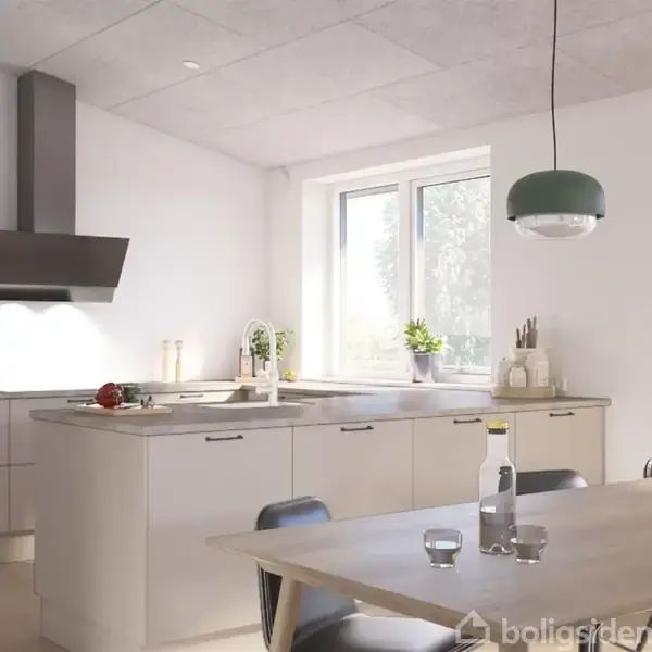 Lysegråt køkken med en stor ø af bordplader i lys træfarve. To grønne lamper hænger over spisebordet. Et vindue giver naturligt lys. Flasker og blomster pynter bordet.