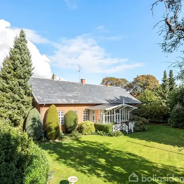 Et rødt murstenshus med sort tag står blandt frodige træer og buske i en velplejet have under en klar blå himmel.