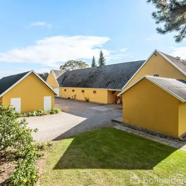 Gulmalede gårdbygninger står stille i solbeskinnet gårdsplads omgivet af græsområder og træer. Blå himmel og spredte skyer danner baggrund.