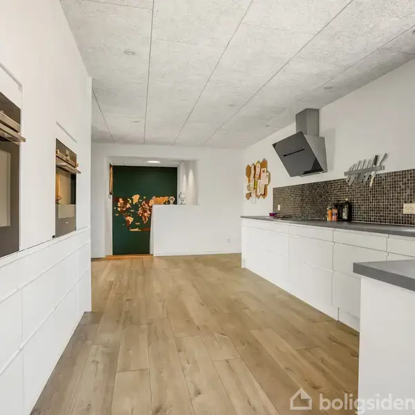 Køkken med hvide skabslåger og integrerede ovne, står tomt. Rummet har et lyst trægulv, mosaikfliser på væggen, grå bordplader og et vindue.