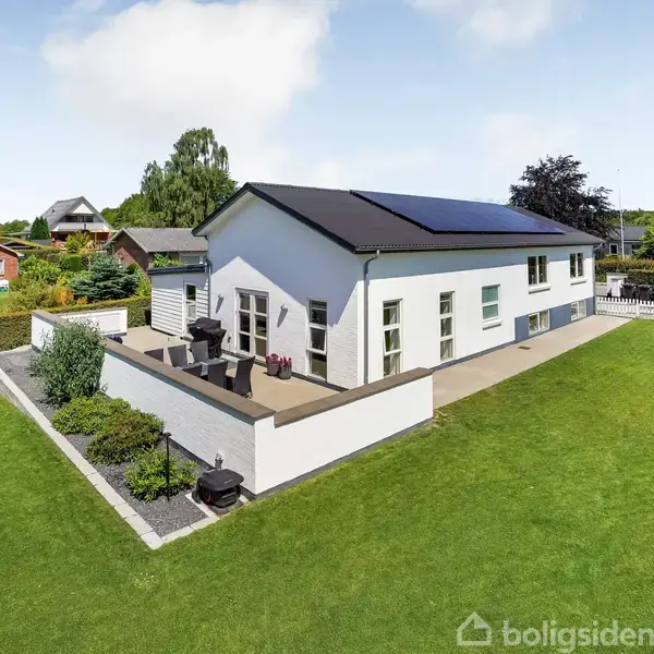 Et hvidt hus med solpaneler på taget står på en grøn græsplæne. Huset har en terrasse med havemøbler. Omgivet af buske og træer i et roligt boligområde.