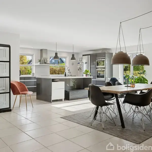 Et moderne køkken-alrum med en stor spisebord i træ og seks sorte stole. Køkkenet har ø med grå skabe, hængende lamper, og store vinduer, der giver naturligt lys.