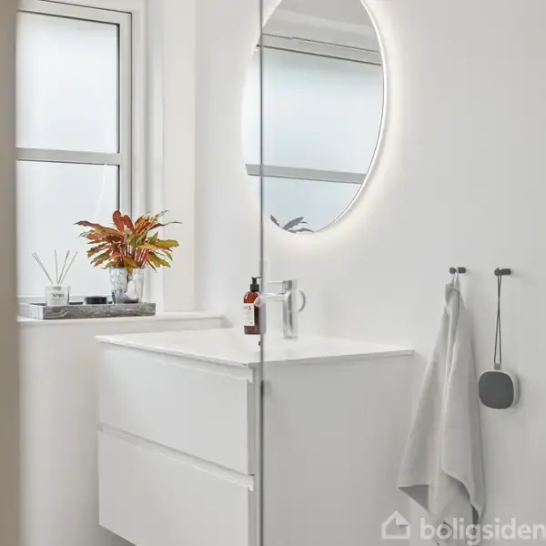 Et rundt spejl hænger over en hvid håndvask i et lyst badeværelse. Ved siden står en glasvase med rødlige blade. Badeværelset er moderne og minimalistisk.