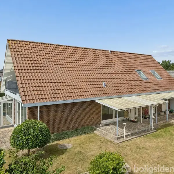 Et rødstenshus med teglstenstag har en terrasseoverdækning og en stor have. Det er omgivet af grønne planter i et boligområde med flere huse synlige i baggrunden.