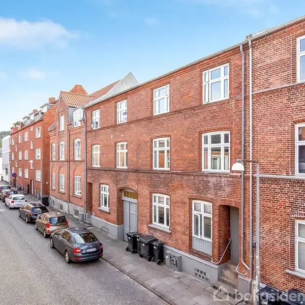Røde murstensbygninger står stille langs en gade med parkerede biler på begge sider, i et fredeligt boligområde under en klar blå himmel.