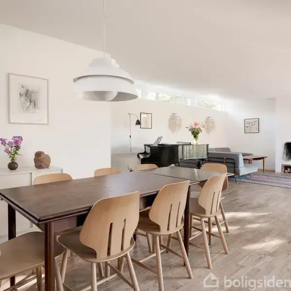 Spisebord omgivet af stole, står på et trægulv. Til venstre er vaser med blomster, til højre er et klaver og en sofa. Væggene har minimalistisk kunst.