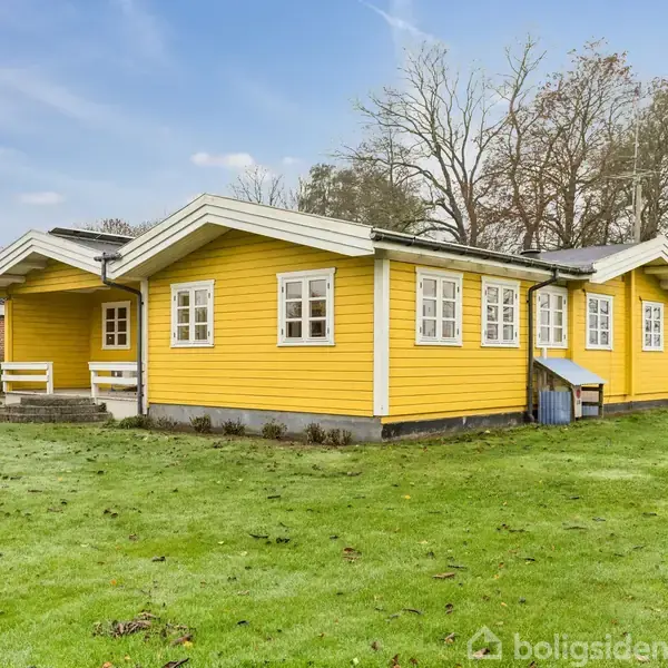 En gul træhytte med hvidmalede vinduesrammer står på en grøn græsplæne. Hytten har en tilbygning og flere vinduer. I baggrunden ses træer og en lille hvid struktur.