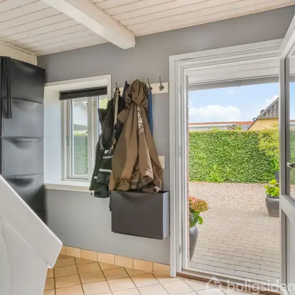 Dør åbner ud til en terrasse med brosten og grønne planter i baggrunden. Jakker hænger ved siden af en trappe i et rum med lyse fliser og hvidt loft.