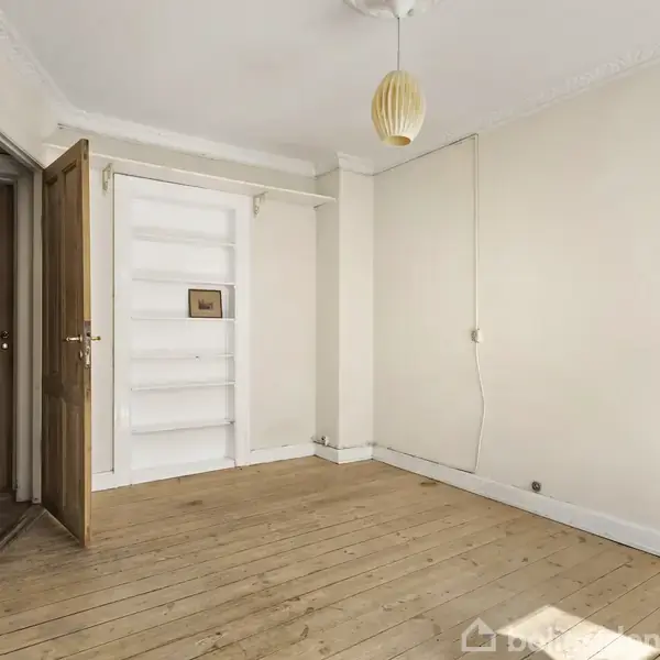 Et tomt rum med trægulv og hvidmalede vægge. En åben dør ses til venstre, og der er en indbygget reol ved siden af døren. En enkel loftlampe hænger ned fra loftet.
