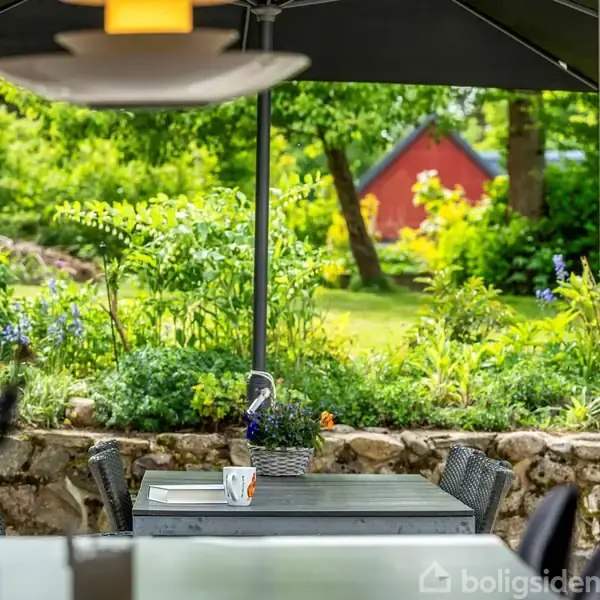 En terrasse med et bord og stole står under en parasol. En kaffekop hviler på bordet. Bagved er en grøn have fyldt med planter og et rødt hus.