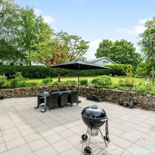 En kuglegrill står på en flisebelagt terrasse ved siden af en spisearrangement med parasol. I baggrunden er en frodig have med grønne træer og buske.