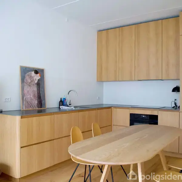 Et træspisebord med stole står i et moderne køkken med lyse træskabe og en mørk bordplade. En åben reol og kunst mod væggen skaber en minimalistisk atmosfære.