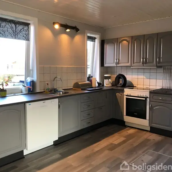 Et køkken med grå skabe og skuffer. En opvaskemaskine er indbygget under en sort bordplade. Der er en hvid komfur og en vask. Lysegrå fliser pryder væggen over bordpladen.