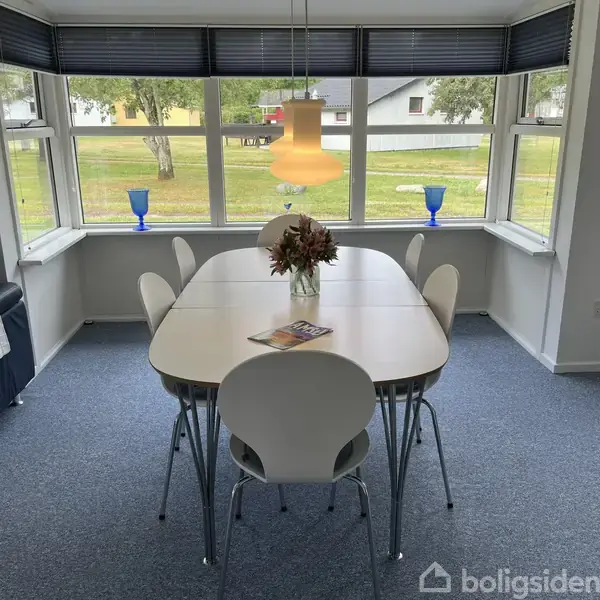 Et spisebord med seks hvide stole står i et lyst rum med mange vinduer. En buket blomster og et blad pryder bordet. Udsigt til en grøn have.