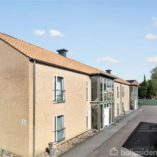 En to-etagers bygning med gul murstensfacade og rødt tegltag står stille ved siden af en belagt indkørsel. Bygningen har store vinduer og ligger i et beboelsesområde.