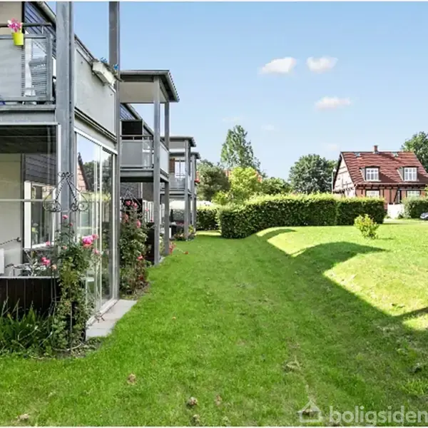 Lejligheder med altaner står i række på grøn græsplæne. Sollys kaster skygger på græsset, mens buske og træer omkranser området. I baggrunden ses et rødt hus.