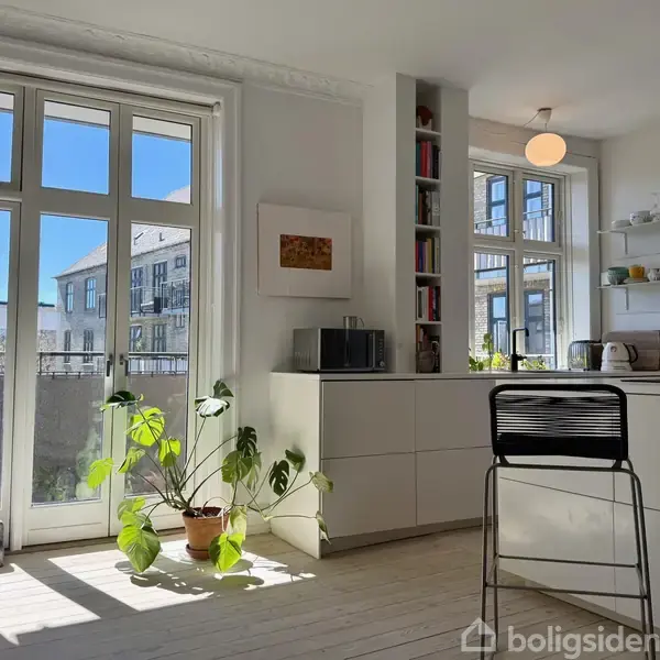 Stue med lyse trægulve indeholder en sofa, en høj plante og et moderne køkkenhjørne med høje barstole. Store vinduer giver udsigt til en bymæssig baggrund og lyseblå himmel.
