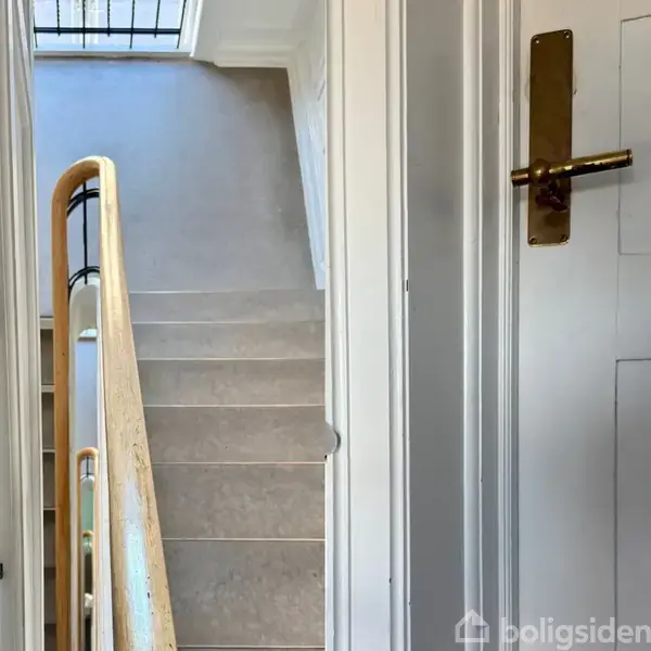 Trappe med beige trin, trægelænder stiger opad i en lys trappeopgang. Dør til højre med gyldent håndtag. Vinduet øverst i trappeopgangen slipper lys ind.