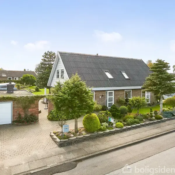 Et hvidt hus med sort tag står i en velplejet have med buske og blomsterbede. Ved siden af huset er en garage. Beliggende i et grønt boligområde.